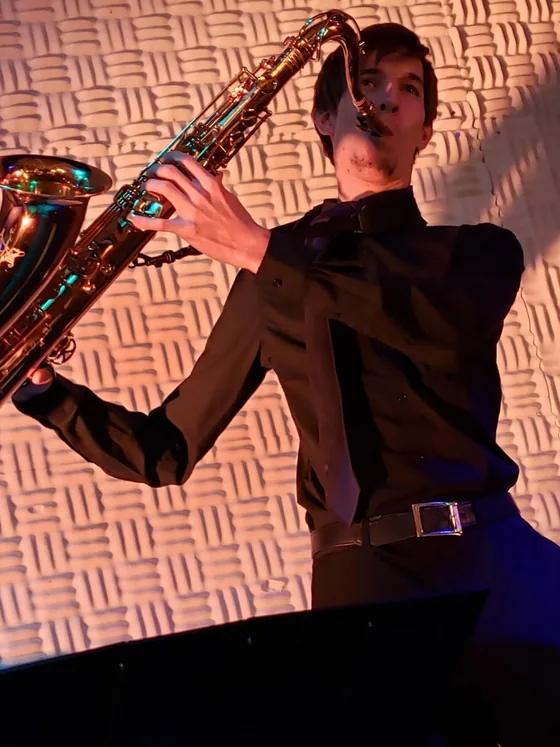 Charlie Suits playing baritone saxophone against a textured wall under warm stage light