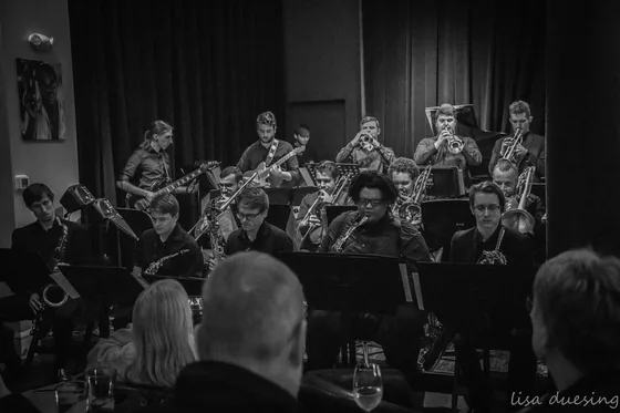 A full big band with a horn section playing to a seated audience, in black and white