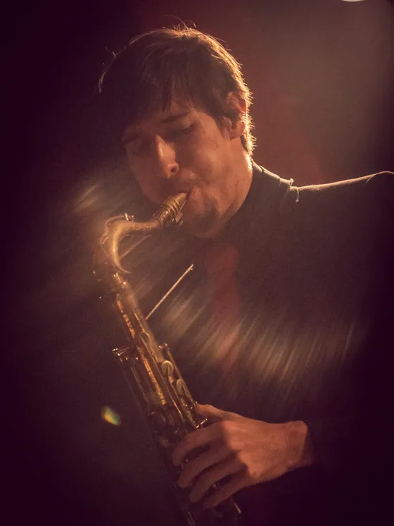 Charlie Suits playing soprano saxophone, lit from behind on a dark stage