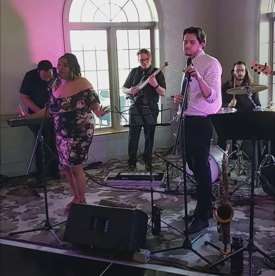 Suits Big Band playing a wedding reception: vocalist, saxophone, guitar, bass and drums in a ballroom with arched windows and uplighting